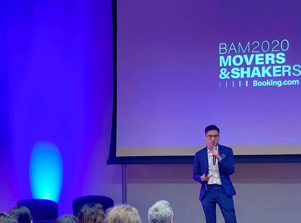 Tim Dương diễn thuyết trên sân khấu tại sự kiện Booking.com BAM 2020 Movers & Shakers, trước khán giả và màn hình mang thương hiệu Booking.com